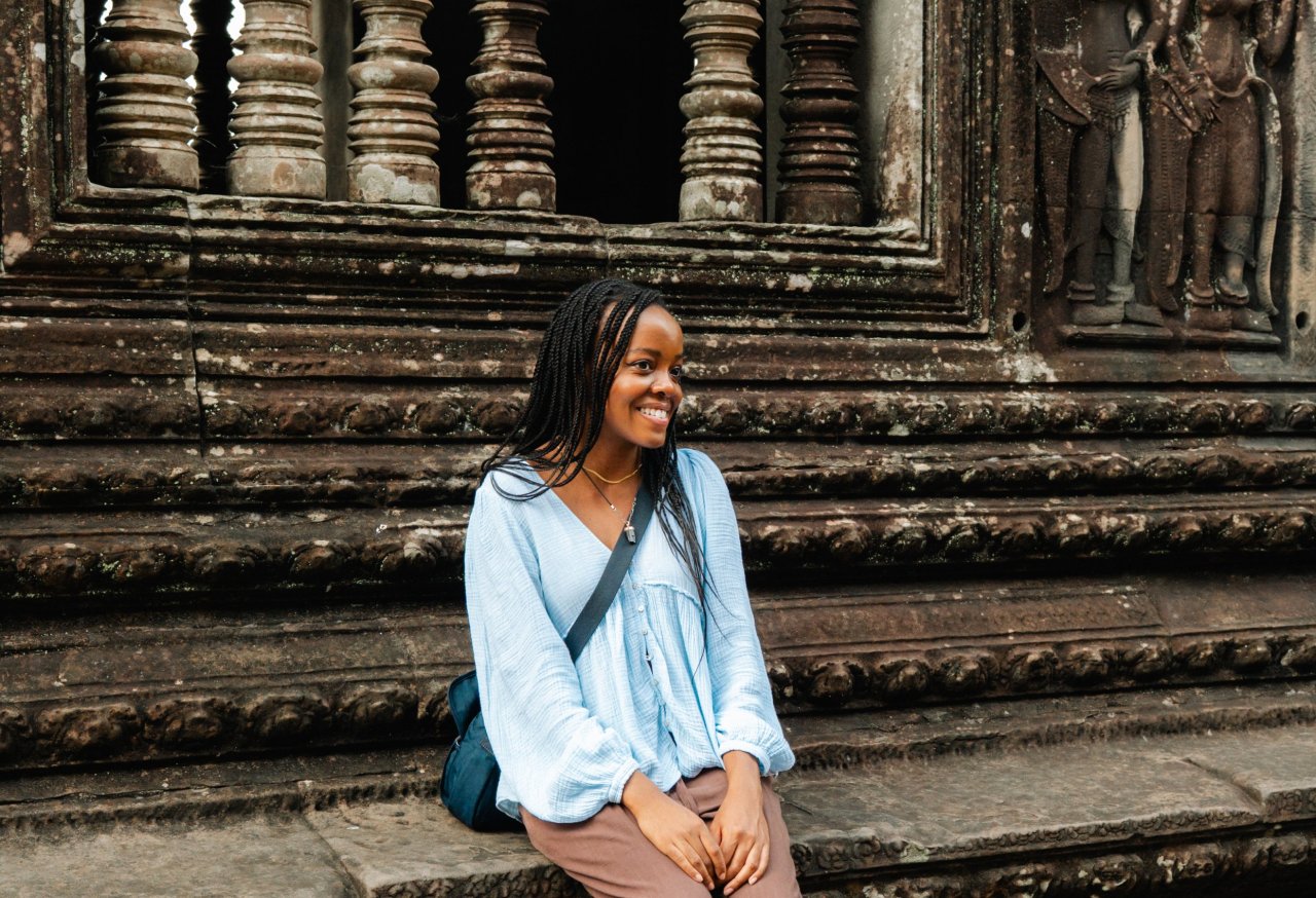 A smiling young traveler sitting on a stone ledge outside the temple walls, framed by carved pillars and stone artwork. Backpacking in Angkor Wat.