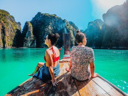 Two girls sat at the edge of a long tail boat admiring the view of bright turquoise water and surrounding cliffs in Koh Phi Phi, Thailand 